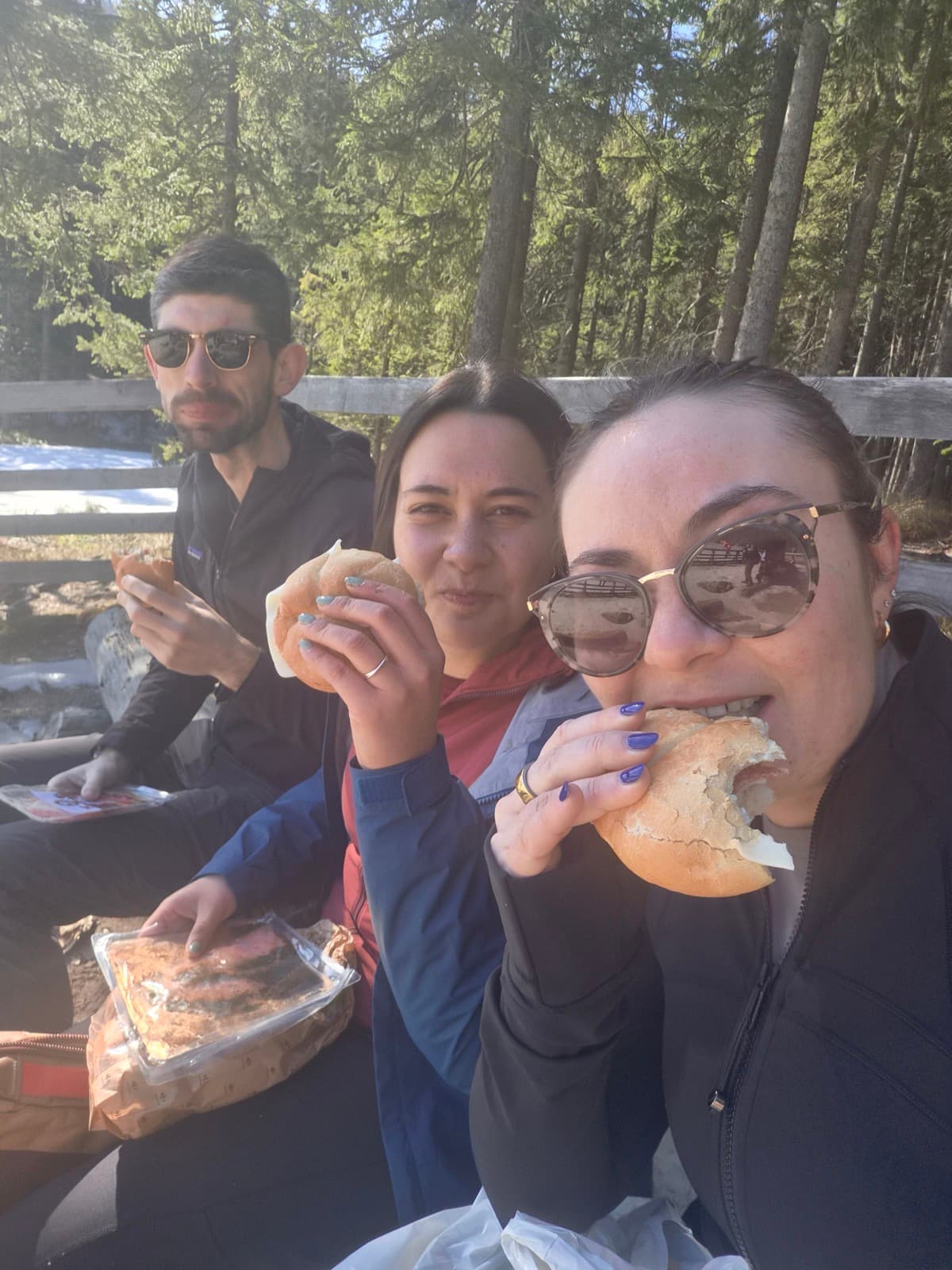 Three people sitting on a snowy bank eating bread rolls by a frozen lake