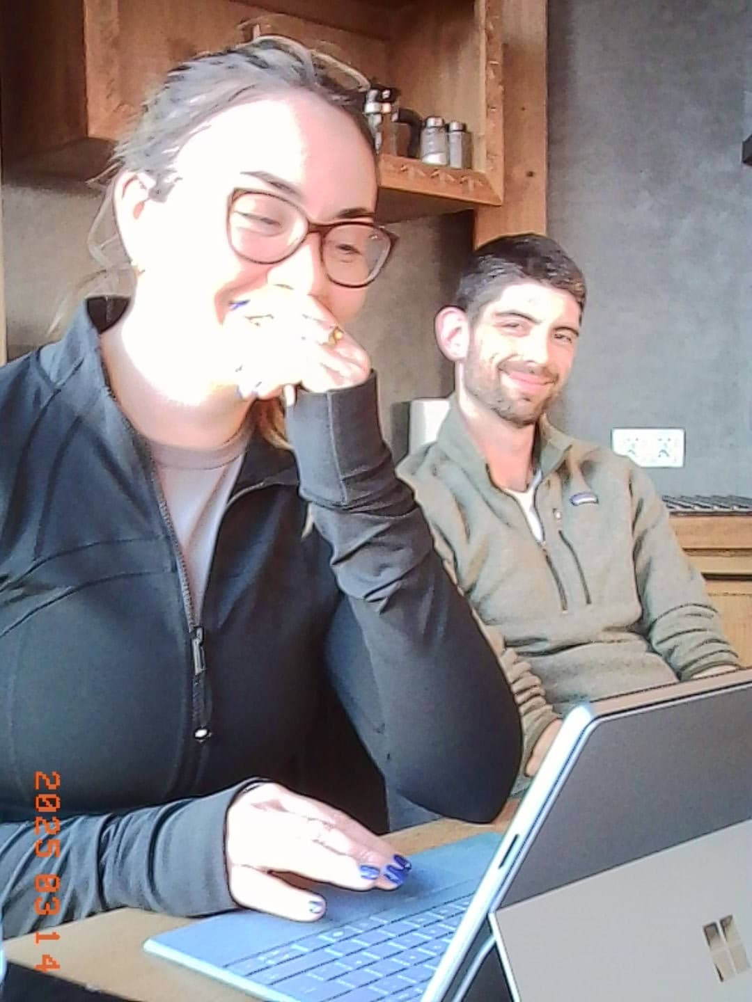 Two people working on laptops at a wooden table by a window